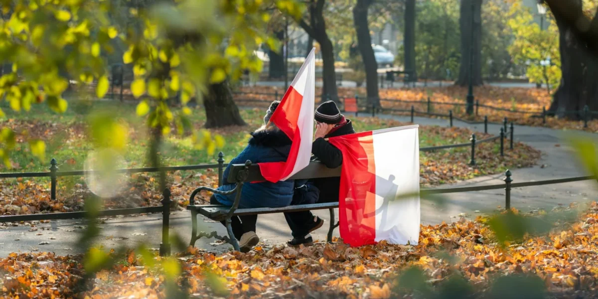 A couple sits on a park bench waving Polish flags, surrounded by autumn leaves in Kraków, Poland.