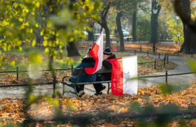 A couple sits on a park bench waving Polish flags, surrounded by autumn leaves in Kraków, Poland.