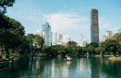 Cityscape view of Bangkok's skyline reflecting in a peaceful park lake under a bright blue sky.