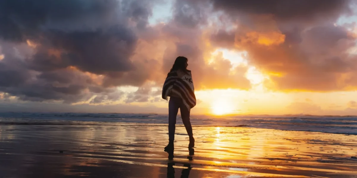 A woman wrapped in a shawl walks on a reflective beach at sunset, creating a moody silhouette against the vibrant sky.