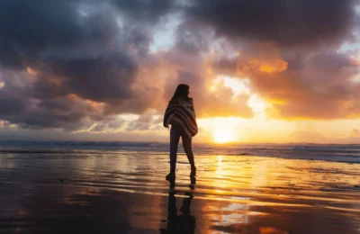 A woman wrapped in a shawl walks on a reflective beach at sunset, creating a moody silhouette against the vibrant sky.