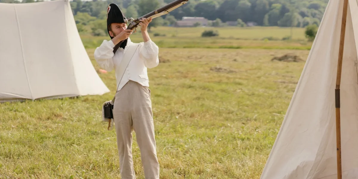 A soldier in historical attire aims a musket during a reenactment on a grassy battlefield.