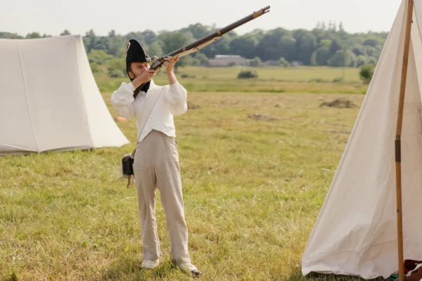 A soldier in historical attire aims a musket during a reenactment on a grassy battlefield.