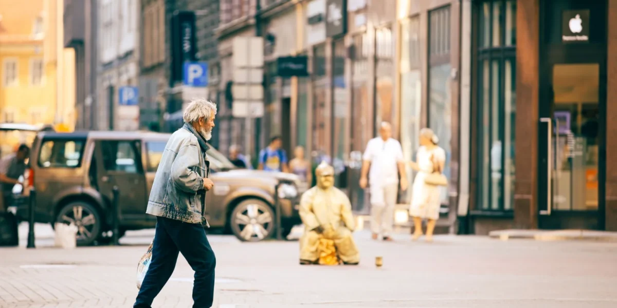 An elderly man in a denim jacket walks past storefronts and parked cars on a bustling city street.
