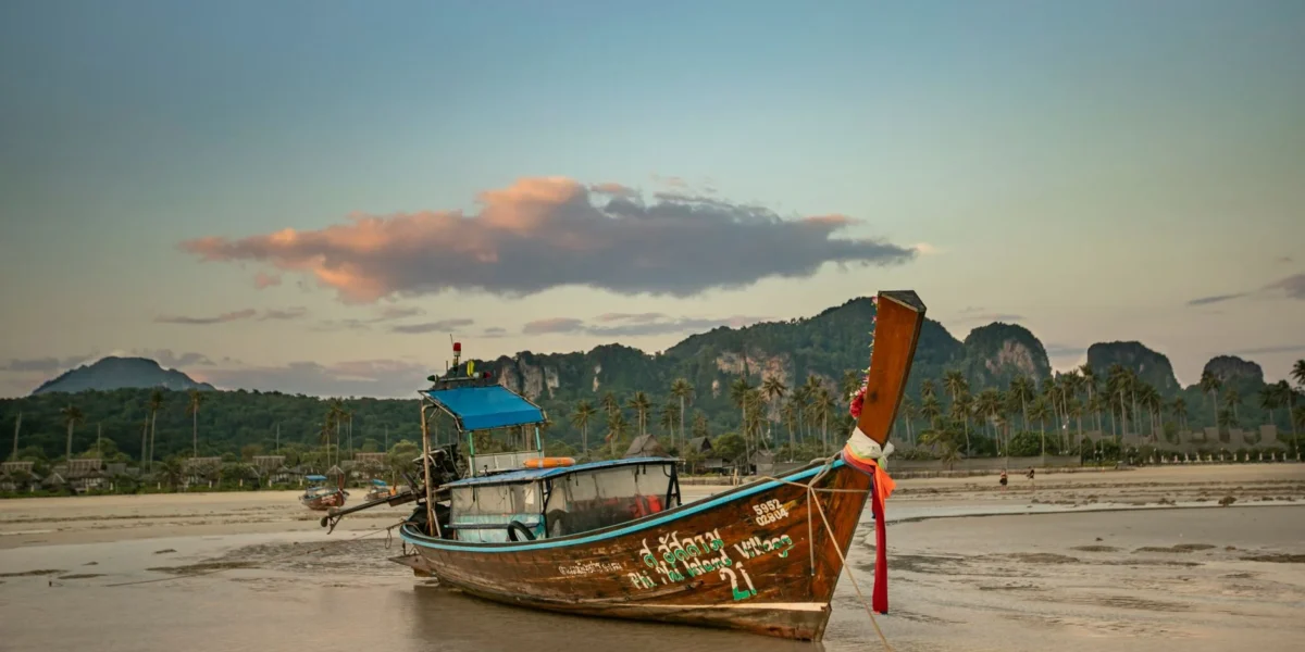 A moored Thai longtail boat on a tranquil beach at sunset with mountains and palm trees in view.