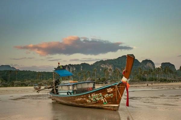 A moored Thai longtail boat on a tranquil beach at sunset with mountains and palm trees in view.